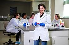 Arwa Hadid in a white lab coat and blue gloves in a Clinical and Diagnostic Sciences lab with other students seated behind her.